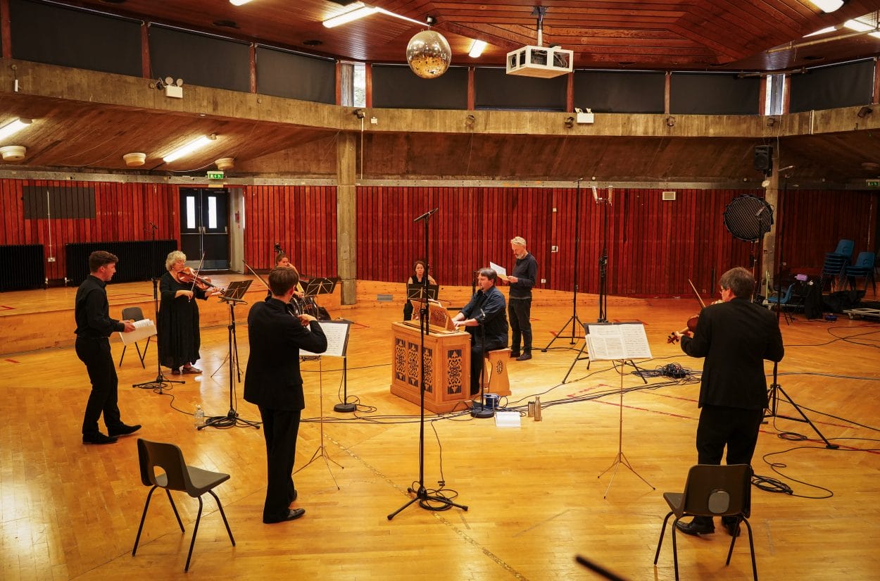 Orchestra players perform in a large, wood-panelled concert hall. A harpsichord player is seated in the centre, surrounded by violinists and a vocalist, with music stands and microphones set up around them.