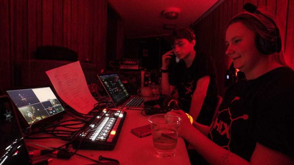 Two students operating video and sound equipment in a dimly lit room with red lighting. One student is sitting, wearing headphones, and holding a glass, while the other, also wearing headphones, is looking at multiple screens and control panels on a desk.