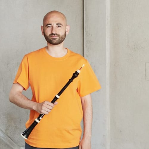 Leo Duarte wears an orange t-shirt and stands in front of a white background. He holds an Oboe in his right hand.