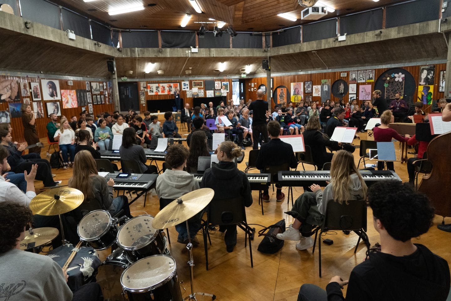 A diverse group of musicians and singers gather in a spacious, wood-paneled room. Some play instruments like drums and keyboards, while others are seated and standing. The walls are adorned with colourful artwork and posters.