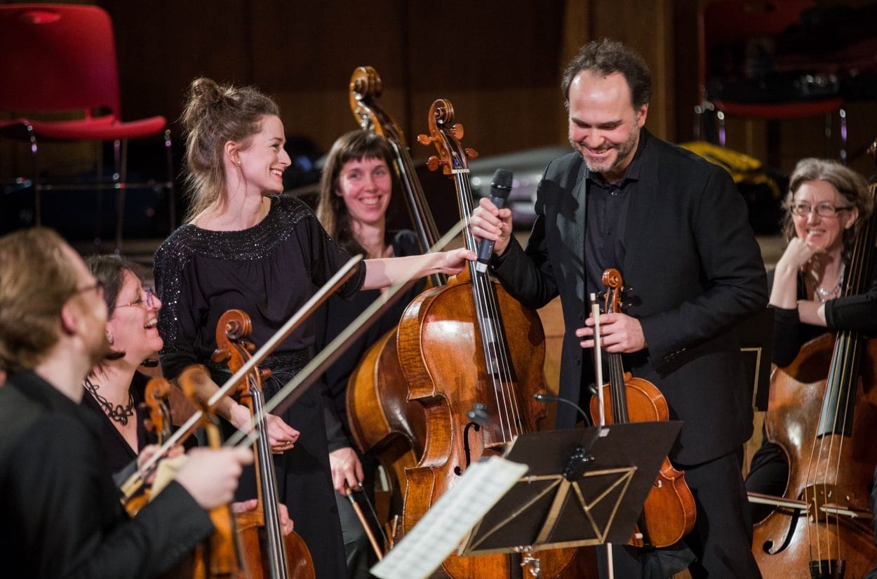 A group of smiling musicians in an orchestra, with a woman in black holding a microphone and passing it to a man. They are surrounded by cellists, and music stands with sheet music are visible.