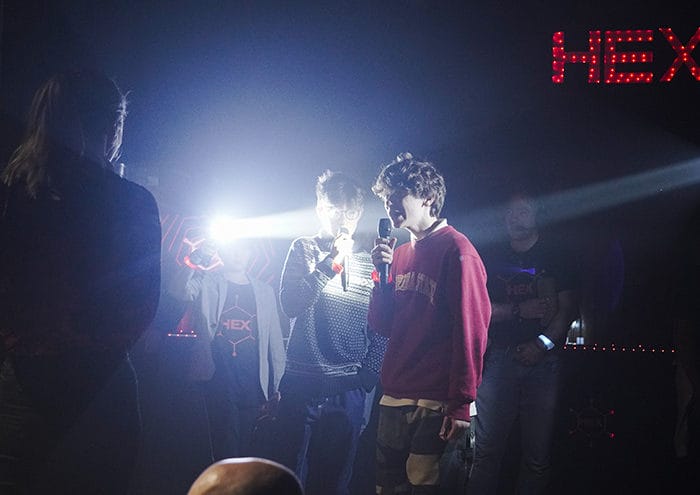 People stand in a dimly lit room with microphones, illuminated by bright lights. The background displays the word "HEX" in red.