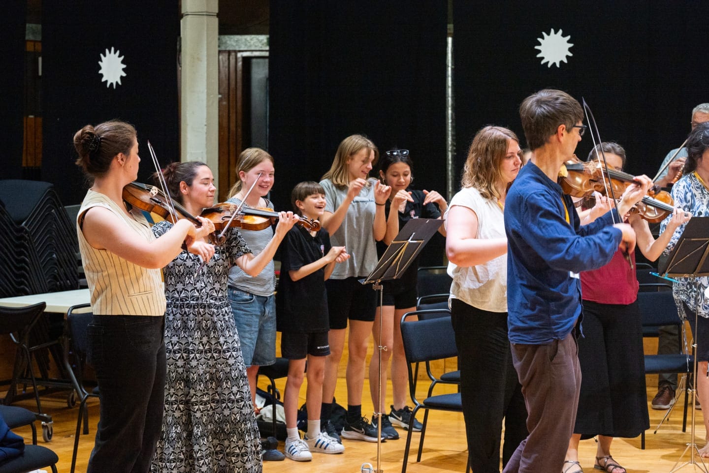 A group of school students are standing in the middle of the violin section of the OAE during a rehearsal.