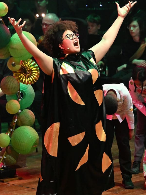 Actress Simon Ibbott-Browne as The Fairy Queen - she wears a black dress with large gold semi-circles and is raising her arms above her head as she sings.