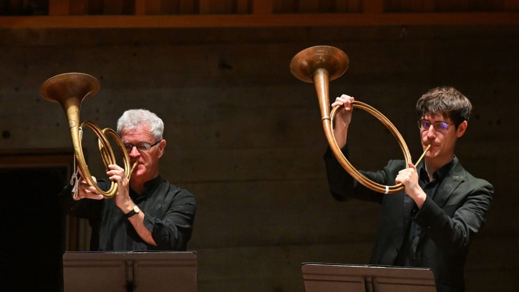 Two horn players from the OAE performing Handel's Solomon - the bells of the instrument are lifted high in the air above the players' heads.